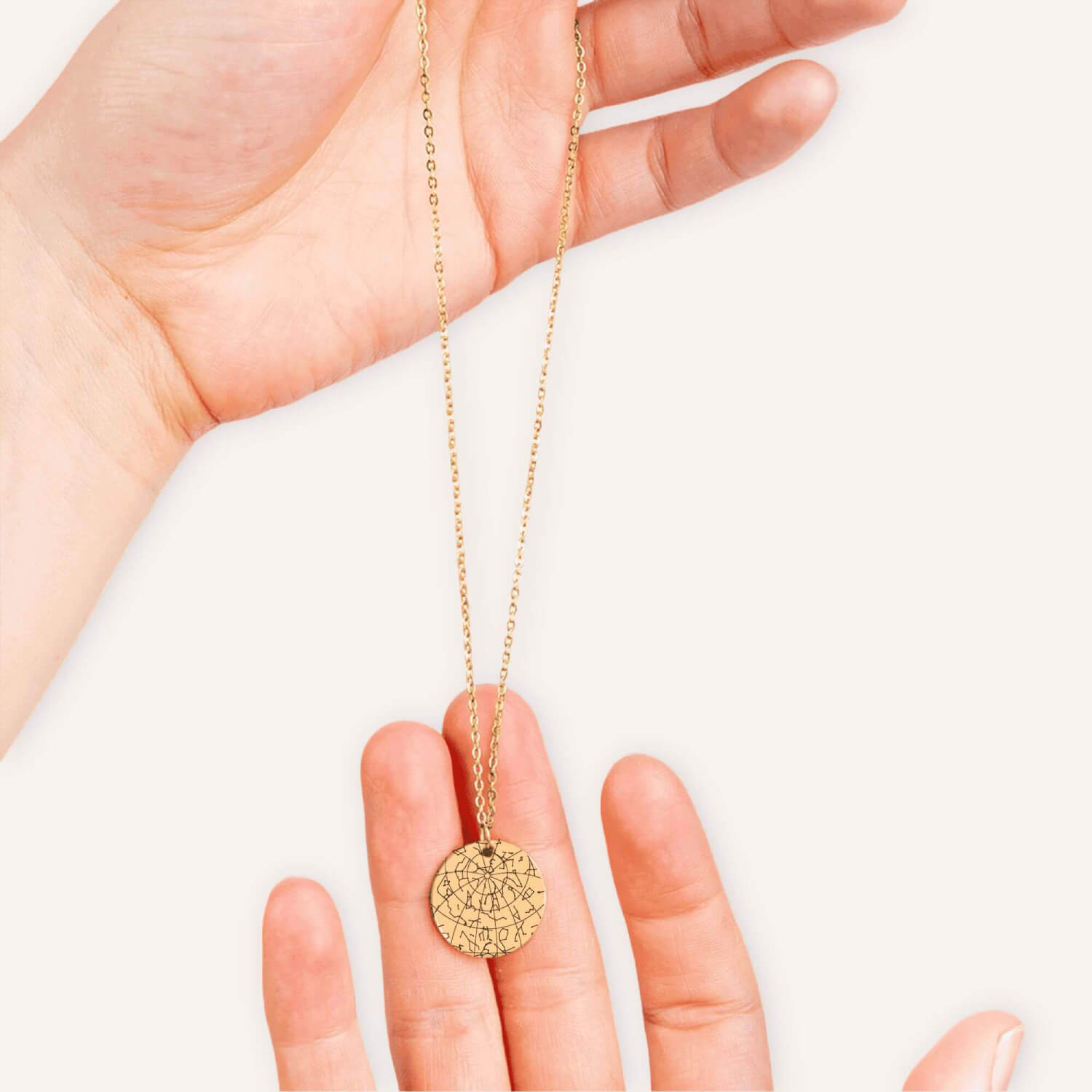 Hand holding a gold necklace with a coin pendent engraved with a custom star map in a natural color background.