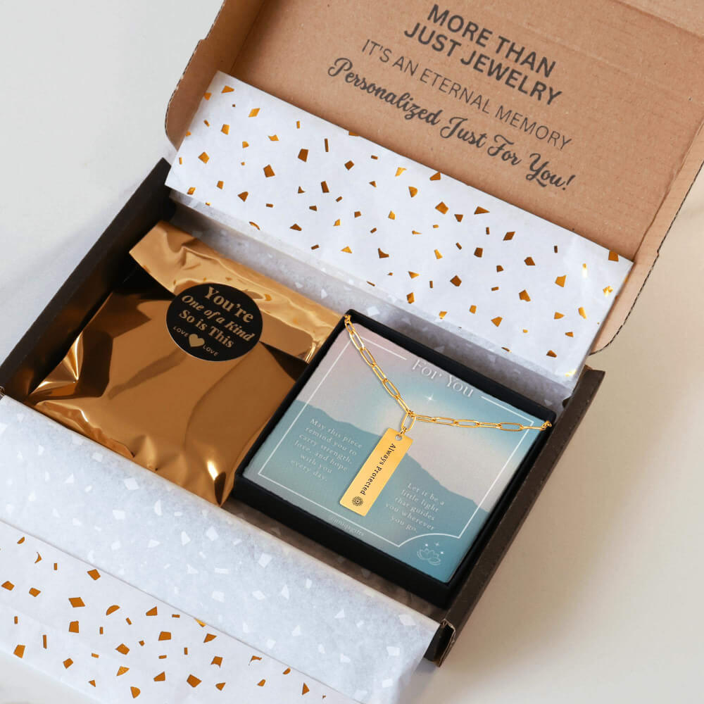 Personalized gold paperclip necklace with a vertical bar pendant engraved with a unique inspiration mantra, packaging in a gift box with a greeting card and gold confetti paper, held by hands against a neutral background.