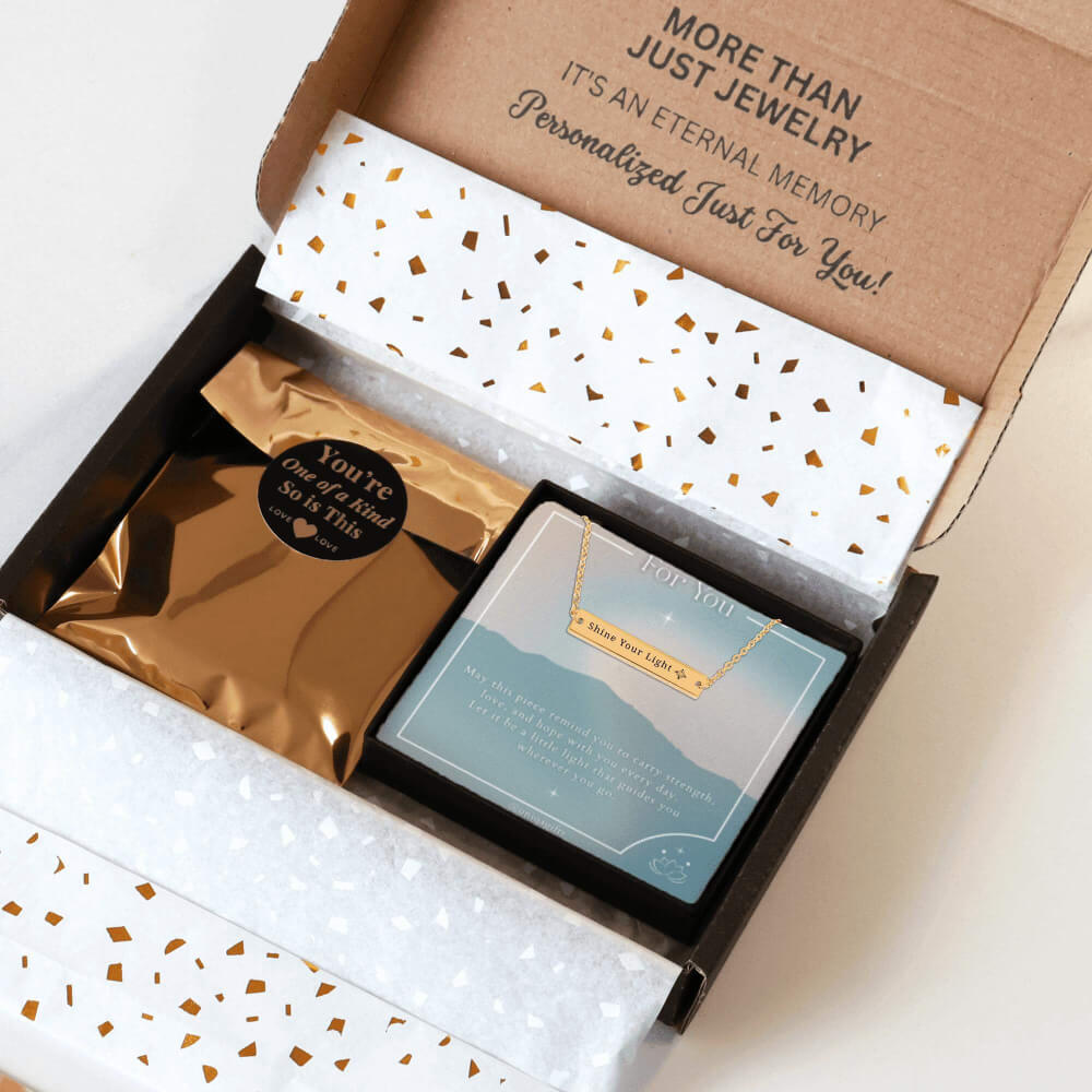 Personalized gold necklace with a bar pendant engraved with a unique inspiration mantra, packaging in a gift box with a greeting card and gold confetti paper, held by hands against a neutral background.