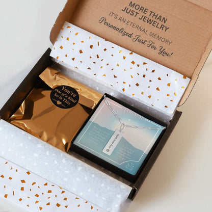 Personalized silver paperclip necklace with a vertical bar pendant engraved with a unique inspiration mantra, packaging in a gift box with a greeting card and gold confetti paper, held by hands against a neutral background.