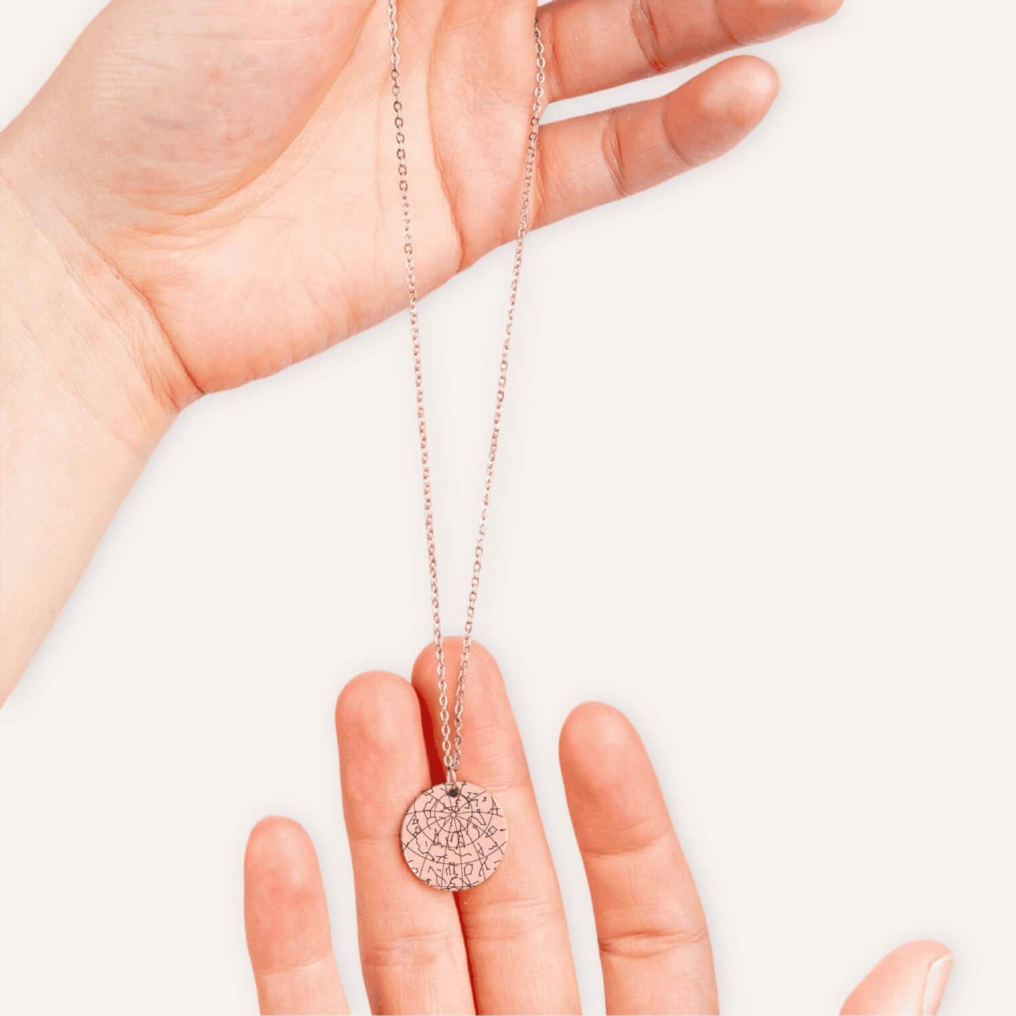 Hand holding a rose gold necklace with a coin pendent engraved with a custom star map in a natural color background.