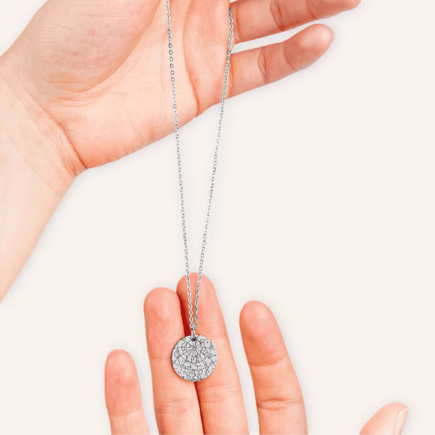 Hand holding a silver necklace with a coin pendent engraved with a custom star map in a natural color background.