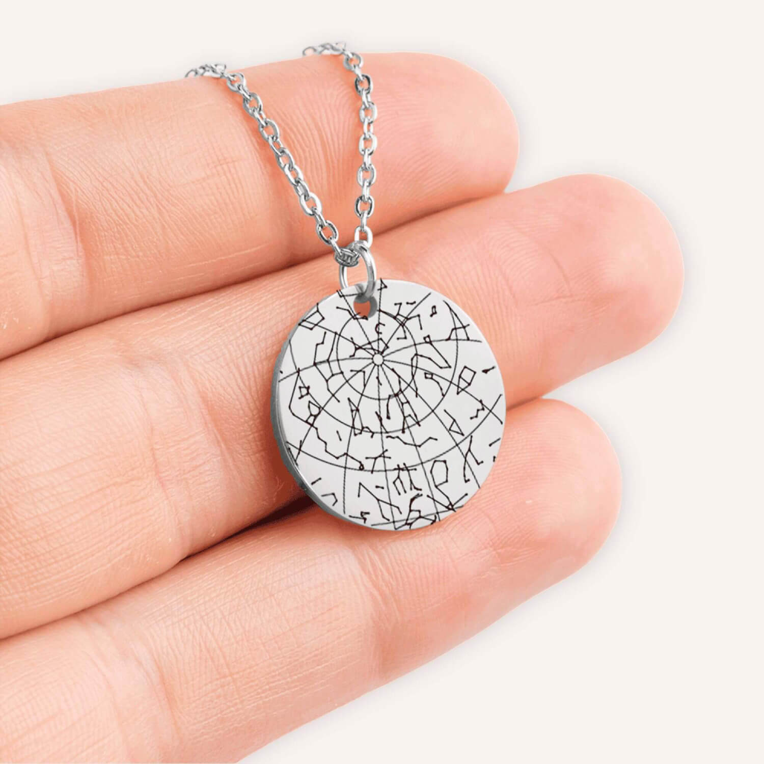 Hand holding a silver coin pendent engraved with a custom star map in a natural color background.