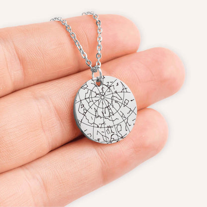 Hand holding a silver coin pendent engraved with a custom star map in a natural color background.