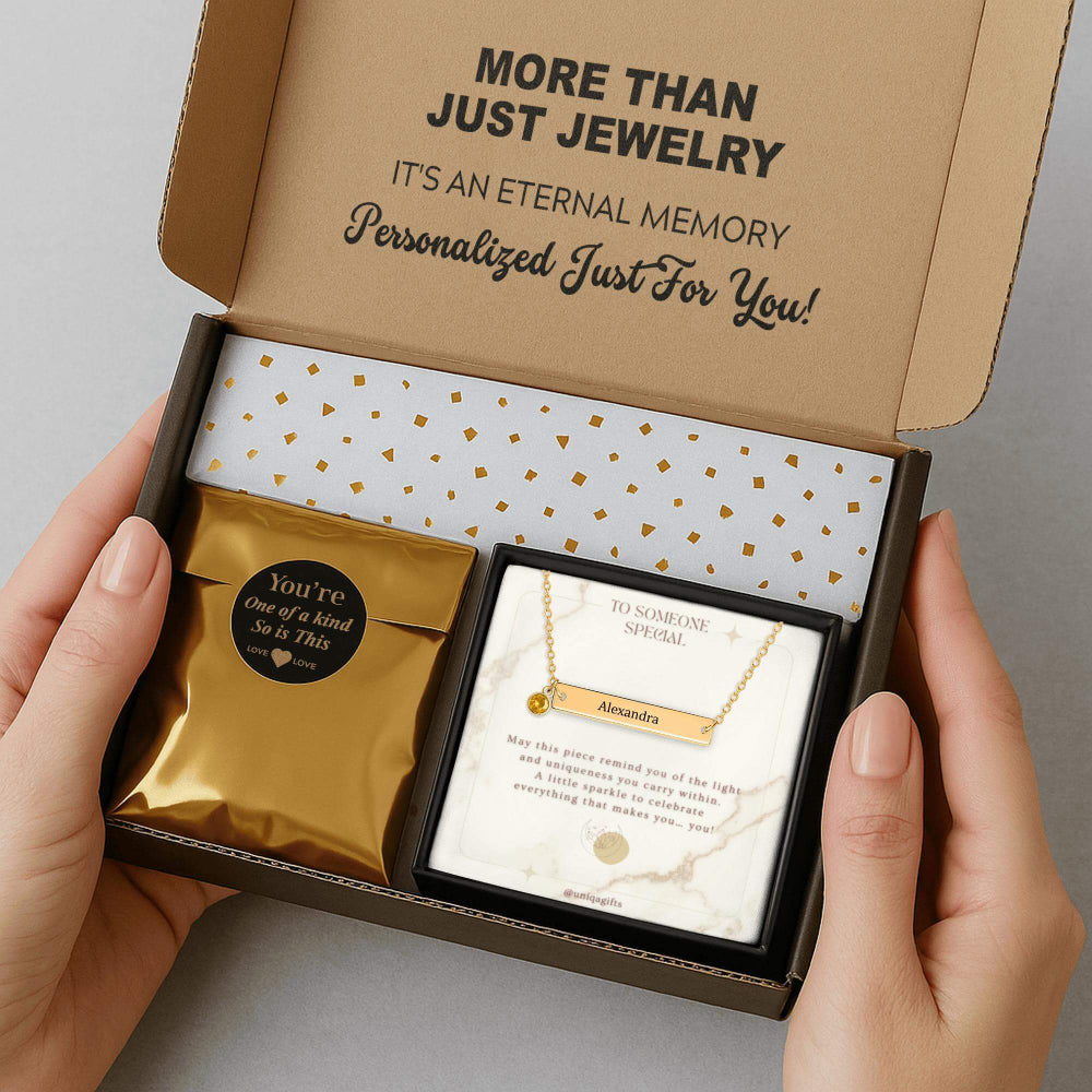 Personalized gold bar necklace with a birthstone, packaging in a gift box with a greeting card and gold confetti paper, held by hands against a neutral background.