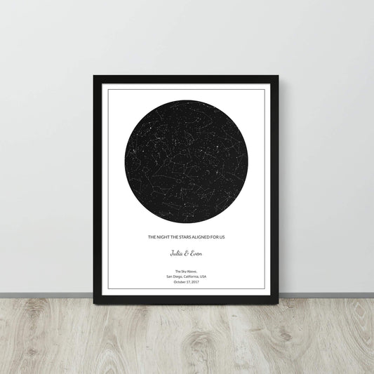 Black and white custom star map poster in a black frame leaning against a light-colored wall on a light wood floor.