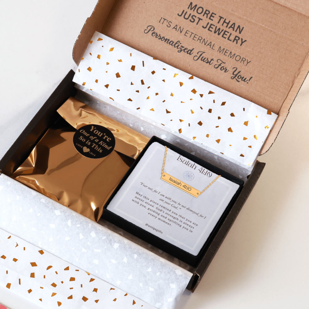 Personalized gold necklace with a bar pendant engraved with an inspirational bible verse, packaging in a gift box with a greeting card and gold confetti paper, held by hands against a neutral background.