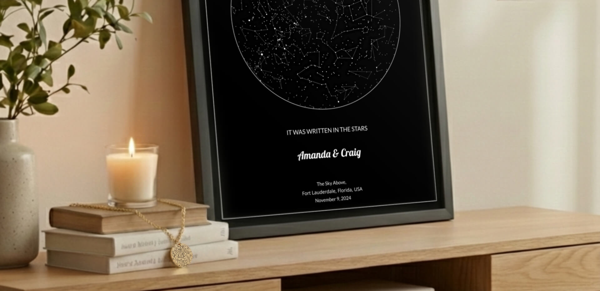 Framed star map poster with personalized text and a star map necklace hanging on books on a table in a warm home setting.